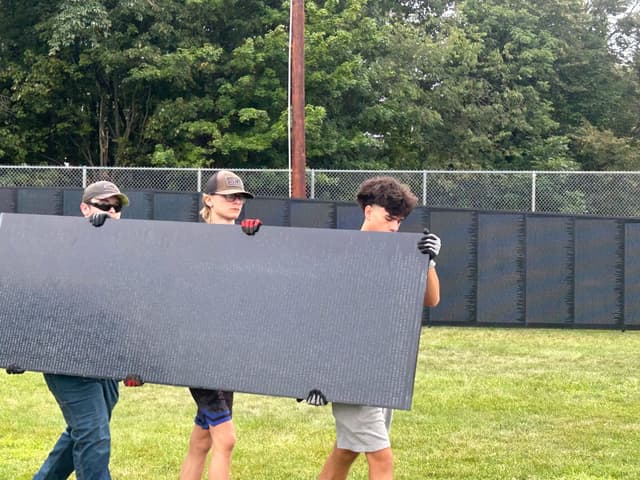 Three volunteers in work gloves carry a panel past the standing Wall
