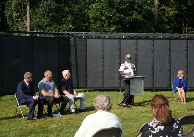 A speaker at the podium during a ceremony, the Wall standing behind