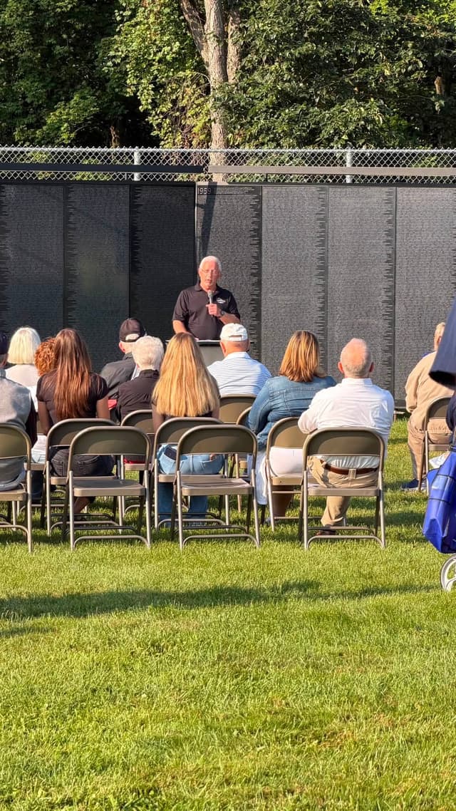 A speaker addresses the seated crowd, the Wall behind him